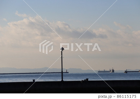 影で黒く見える海岸から青い海面の海と突堤と灯台と漁船とタンカーと霞む山と雲と薄い色の青空の風景 影で黒く見える海岸から青い海面の海と突堤と灯台と漁船とタンカーと霞む山と雲と薄い色の青空の風景 86115175