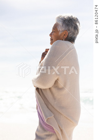 Side view of biracial senior woman with short white hair wearing shrug at beach on sunny day 86120474