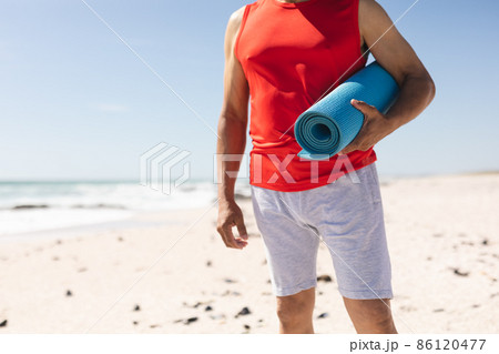 Midsection of retired senior biracial man holding rolled yoga mat at beach on sunny day 86120477
