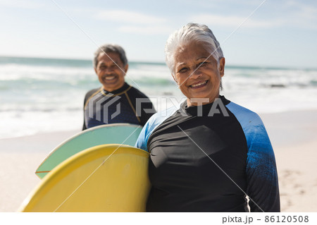 Portrait of smiling biracial senior woman and man carrying surfboards at beach on sunny day Portrait of smiling biracial senior woman and man carrying surfboards at beach on sunny day 86120508