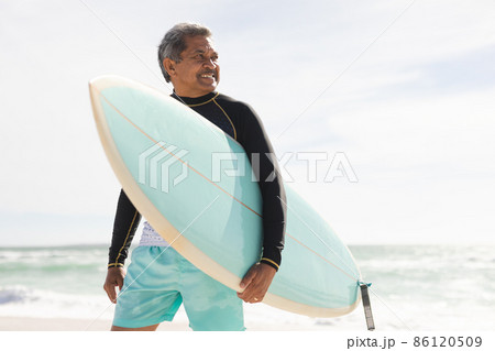 Smiling biracial senior man carrying surfboard looking away at beach against sky during sunny day 86120509