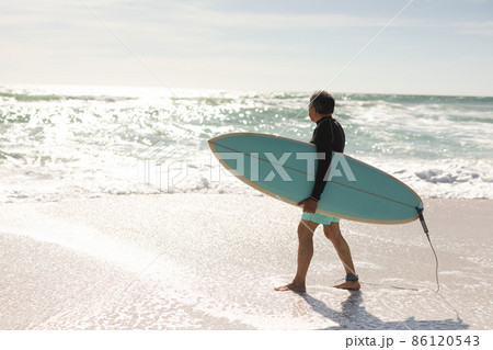 Biracial senior man carrying surfboard wading on shore looking at waves splashing during sunny day 86120543