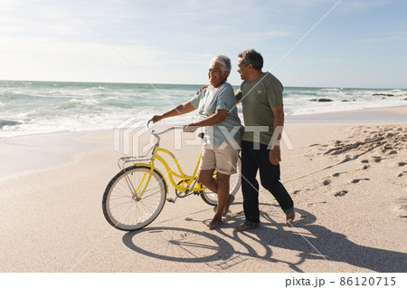 Biracial senior man walking and talking with woman wheeling bicycle at sunny beach against sky 86120715