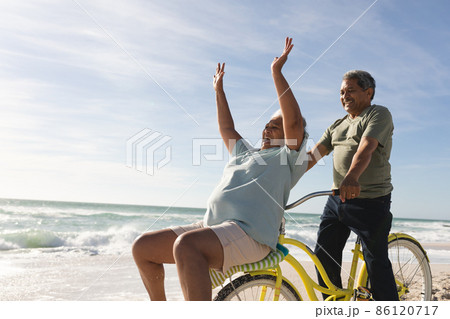 Cheerful biracial senior woman cheering while man riding bicycle at beach on sunny day 86120717