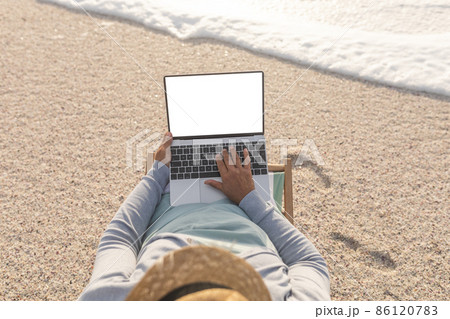 High angle view of senior biracial woman using blank screen laptop with copy space on shore at beach High angle view of senior biracial woman using blank screen laptop with copy space on shore at beach 86120783