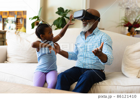 African american girl making her grandfather wear vr headset in living room at home 86121410