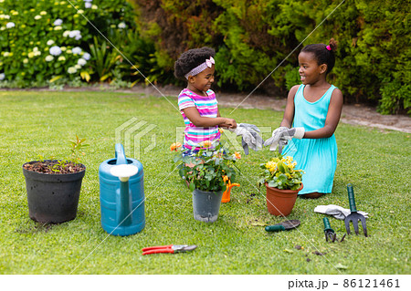 Playful smiling african american sisters kneeling while wearing gloves by plants in garden 86121461