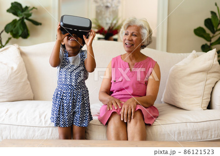 African american girl wearing vr headset and grandmother smiling sitting on the couch African american girl wearing vr headset and grandmother smiling sitting on the couch 86121523