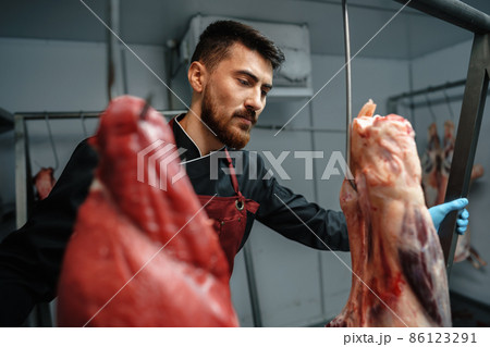 Male butcher standing by meat hanging on hook in meat shop 86123291