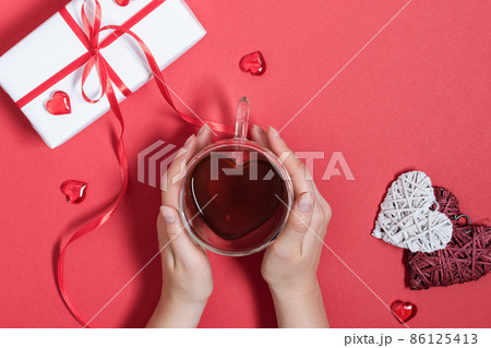 Woman hands holding tea cup in a heart shaped with red hearts decoration on red table top view. 86125413