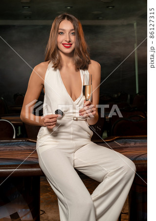 Successful young woman with betting chips and glass of champagne near poker table in gambling establishment 86127315