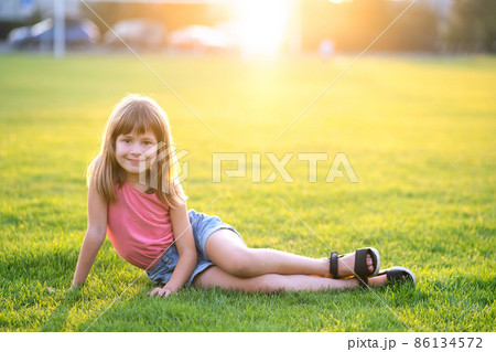 Young happy child girl resting while sitting on green grass lawn on warm summer day. Young happy child girl resting while sitting on green grass lawn on warm summer day. 86134572