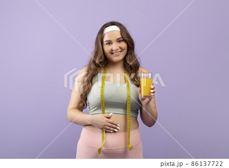 Happy pretty caucasian plus size young woman in sportswear with measuring tape show glass of fresh orange juice Happy pretty caucasian plus size young woman in sportswear with measuring tape show glass of fresh orange juice 86137722