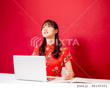 Portrait of a young asian woman in traditional cheongsam qipao dress using laptop and writing on red background. Portrait of a young asian woman in traditional cheongsam qipao dress using laptop and writing on red background. 86140305