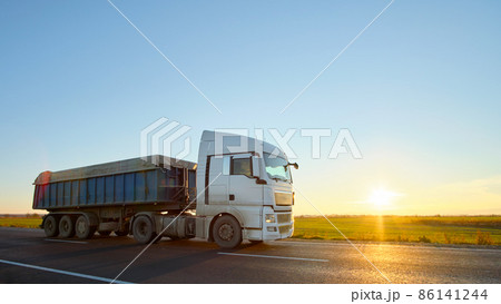 Semi-truck with tipping cargo trailer transporting sand from quarry driving on highway hauling goods in evening. Delivery transportation and logistics concept Semi-truck with tipping cargo trailer transporting sand from quarry driving on highway hauling goods in evening. Delivery transportation and logistics concept 86141244