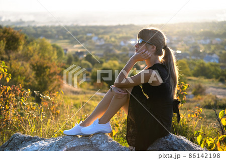 Young pretty woman in black short summer dress sitting on a rock relaxing outdoors at sunset. Fashionable female enjoying warm evening in nature. 86142198