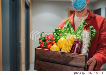 Delivery man in protective mask holding paper bag with food in the entrance. The courier gives the box with fresh vegetables and fruits to the customer Delivery man in protective mask holding paper bag with food in the entrance. The courier gives the box with fresh vegetables and fruits to the customer 86148901