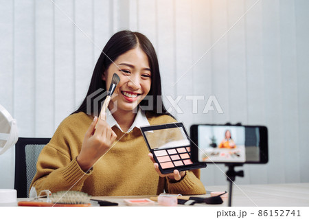 Beautiful asian woman blogger using camera phone recording vlog video live streaming and showing how to make up cosmetics at home. Beautiful asian woman blogger using camera phone recording vlog video live streaming and showing how to make up cosmetics at home. 86152741