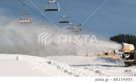 Snow cannon making snow by daytime. Ski lift with skiers is moving in background, skiers and snowboarders drive down the snow covered slope. Snowmaker produces a lot artificial snow Snow cannon making snow by daytime. Ski lift with skiers is moving in background, skiers and snowboarders drive down the snow covered slope. Snowmaker produces a lot artificial snow 86154995