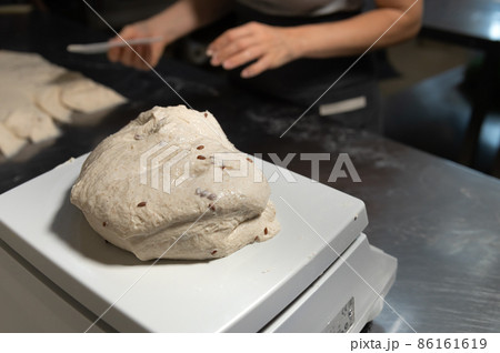 Close-up on a production scale is a portion of bread dough. Hands of a woman baker in defocus in the background. Bread production in a bakery Close-up on a production scale is a portion of bread dough. Hands of a woman baker in defocus in the background. Bread production in a bakery 86161619