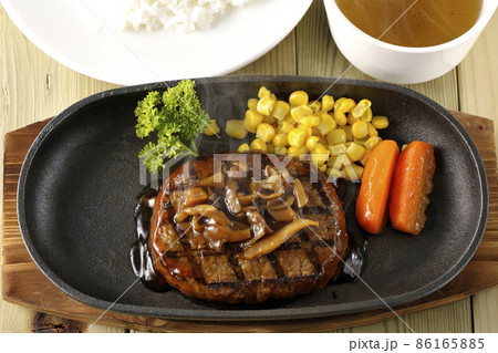 鉄板の上のパセリ、グラッセ、コーン、きのこソースの熱々和風ハンバーグステーキとコンソメスープとライス 86165885