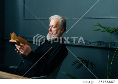 Focused senior gray-haired aged male reading paper book at home sitting at table, relax and rest with literature. Handsome mature adult man reading book at home alone. Focused senior gray-haired aged male reading paper book at home sitting at table, relax and rest with literature. Handsome mature adult man reading book at home alone. 86168887