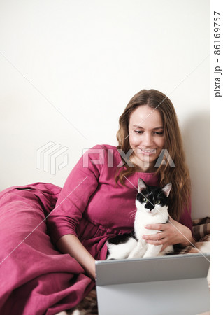 Young millennial woman in pink dress with long hair using a tablet and watcing movies with her black and white tuxedo cat at home. Young millennial woman in pink dress with long hair using a tablet and watcing movies with her black and white tuxedo cat at home. 86169757