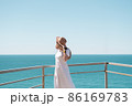 Young woman in white dress and hat stands by the sea. 86169783