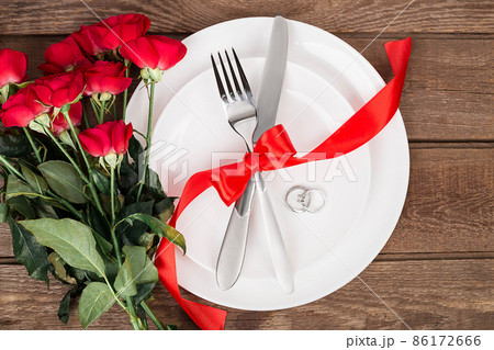 Top view closeup of romantic dinner serving with a bouquet red roses and ring above the white plate Top view closeup of romantic dinner serving with a bouquet red roses and ring above the white plate 86172666