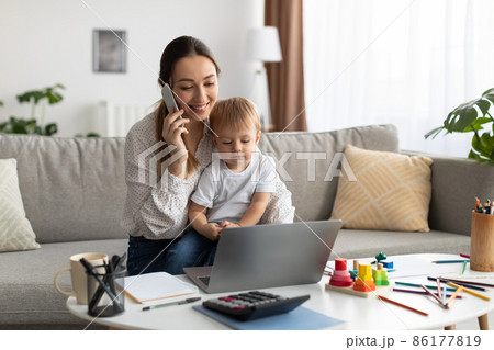 Happy woman talking on cellphone and using laptop with baby sitting on mom's knees, free space Happy woman talking on cellphone and using laptop with baby sitting on mom's knees, free space 86177819
