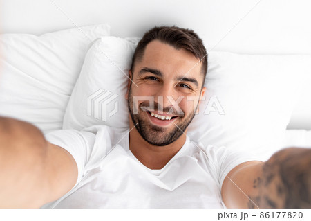 Satisfied happy attractive young caucasian muscular man lies on white bed and makes selfie in bedroom, close up Satisfied happy attractive young caucasian muscular man lies on white bed and makes selfie in bedroom, close up 86177820