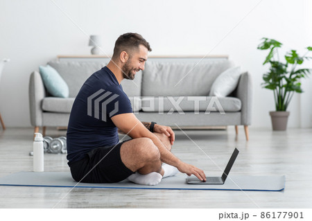 Happy young caucasian muscular guy sits in lotus position on mat and looks at laptop Happy young caucasian muscular guy sits in lotus position on mat and looks at laptop 86177901