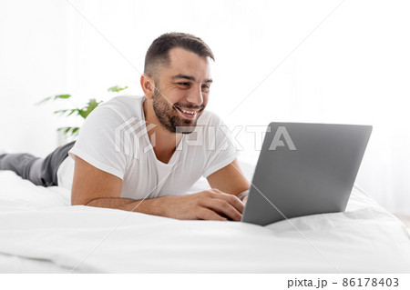 Happy smiling young european muscular man lies on white bed and chatting on computer, watch funny video 86178403