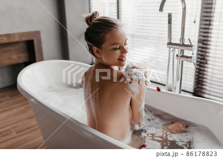 Rear view of pretty lady taking bath and playing with foam, touching shoulder, relaxing in bathroom at home Rear view of pretty lady taking bath and playing with foam, touching shoulder, relaxing in bathroom at home 86178823