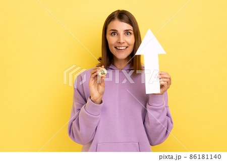 Portrait of young adult attractive dark haired woman showing growth increase of bitcoin, expressing positive emotions, wearing purple hoodie. Indoor studio shot isolated on yellow background. Portrait of young adult attractive dark haired woman showing growth increase of bitcoin, expressing positive emotions, wearing purple hoodie. Indoor studio shot isolated on yellow background. 86181140