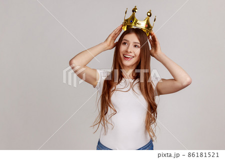 Portrait of happy dark haired woman putting crown on head and smiling, concept of self confidence in success, self-motivation, wearing white T-shirt. Indoor studio shot isolated on gray background. 86181521