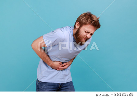 Stomach pain. Side view of bearded man standing and holding his painful belly, feeling bad, suffering diarrhea or constipation, risk of appendicitis. Indoor studio shot isolated on blue background. Stomach pain. Side view of bearded man standing and holding his painful belly, feeling bad, suffering diarrhea or constipation, risk of appendicitis. Indoor studio shot isolated on blue background. 86181539
