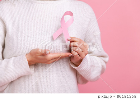 Closeup of unknown female holding pink ribbon, symbol of breast cancer awareness, cancer prevention, wearing white casual style sweater. Indoor studio shot isolated on pink background. 86181558