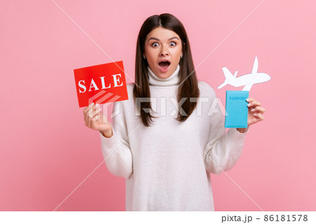 Shocked surprised girl holding passport, paper plane and sale card, vacation abroad for low price, wearing white casual style sweater. Indoor studio shot isolated on pink background. Shocked surprised girl holding passport, paper plane and sale card, vacation abroad for low price, wearing white casual style sweater. Indoor studio shot isolated on pink background. 86181578