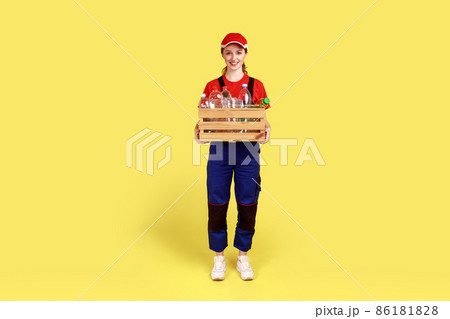 Full length portrait of positive satisfied woman volunteer standing with box full of empty plastic bottles, wearing overalls and red cap. Indoor studio shot isolated on yellow background. Full length portrait of positive satisfied woman volunteer standing with box full of empty plastic bottles, wearing overalls and red cap. Indoor studio shot isolated on yellow background. 86181828