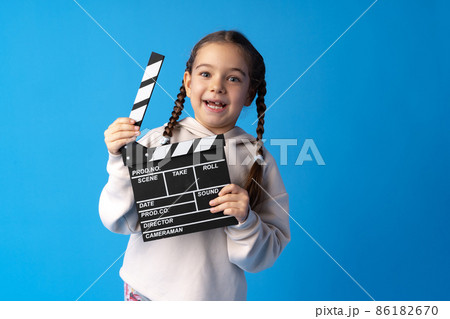Smiling girl holding clapper board against blue background Smiling girl holding clapper board against blue background 86182670