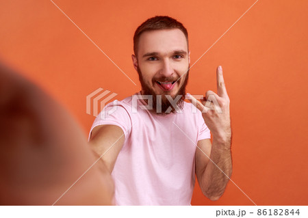 Portrait of handsome man making selfie or streaming, showing rock and roll gesture and tongue out, point of view of photo, wearing pink T-shirt. Indoor studio shot isolated on orange background. Portrait of handsome man making selfie or streaming, showing rock and roll gesture and tongue out, point of view of photo, wearing pink T-shirt. Indoor studio shot isolated on orange background. 86182844