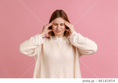 Portrait of unhealthy sick blond woman massaging temples with hands feeling terrible headache, risk of migraine, wearing white sweater. Indoor studio shot isolated on pink background. Portrait of unhealthy sick blond woman massaging temples with hands feeling terrible headache, risk of migraine, wearing white sweater. Indoor studio shot isolated on pink background. 86182854