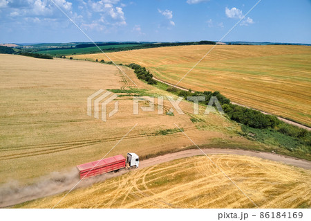 Aerial view of lorry cargo truck driving on dirt road between agricultural wheat fields. Transportation of grain after being harvested by combine harvester during harvesting season 86184169