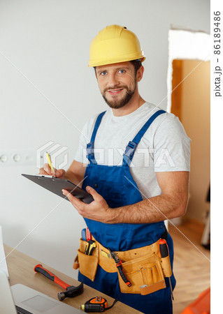 Joyful handyman worker, builder in uniform and hard hat looking at camera, making notes on a clipboard while standing indoors, getting ready for renovation work 86189486
