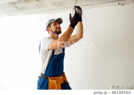 Focused electrician in uniform installing light fitting in new apartment, standing on a ladder Focused electrician in uniform installing light fitting in new apartment, standing on a ladder 86189560