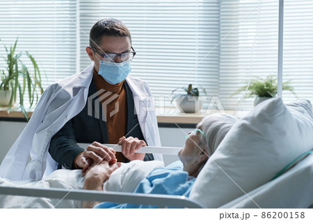 Young son in protective mask visiting his sick senior mother at hospital ward during pandemic Young son in protective mask visiting his sick senior mother at hospital ward during pandemic 86200158