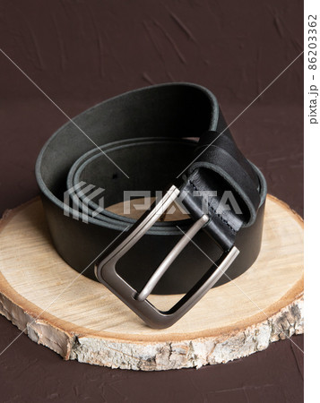 Black fashionable men's leather belt with metal buckle on wood slice on brown background. Male accessory. Front view, vertical photo. 86203362