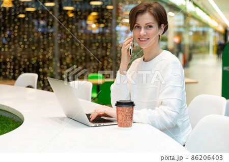 A photo of a positive smiling young woman who sits at the food court of a shopping center at a table A photo of a positive smiling young woman who sits at the food court of a shopping center at a table 86206403