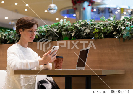 A photo of a young woman who sits at the food court of a shopping center at a table and works at a A photo of a young woman who sits at the food court of a shopping center at a table and works at a 86206411